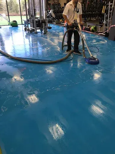 A person uses a floor scrubber to clean a blue, reflective gym floor.