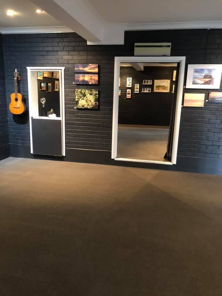 A dark-walled gallery with an acoustic guitar, framed art, and a doorway leading into another exhibit room.