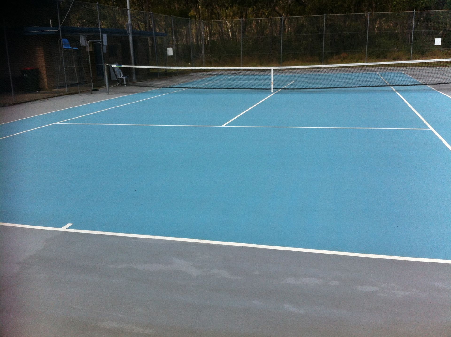 An empty blue outdoor tennis court with white lines and a net, surrounded by a chain-link fence.