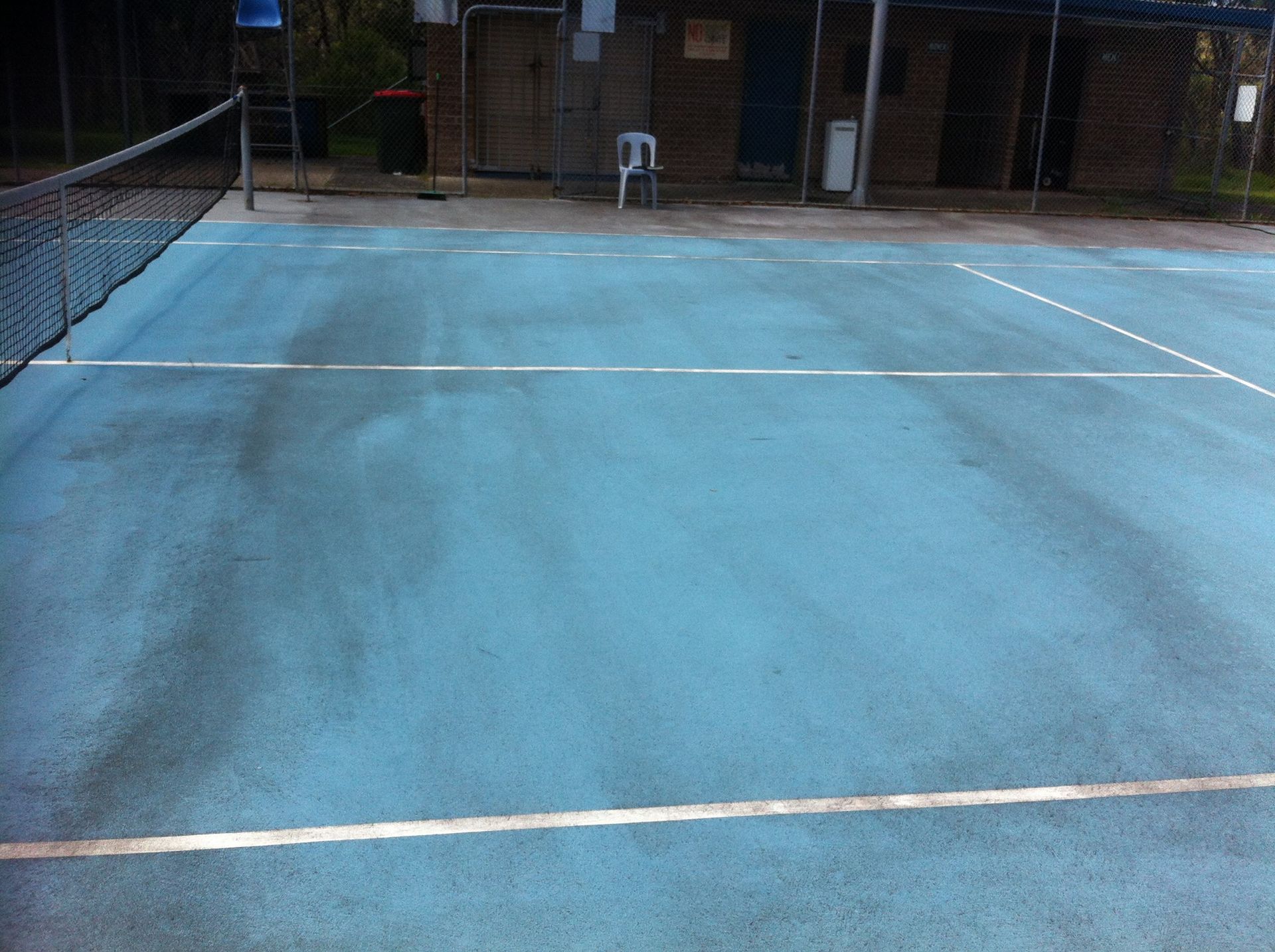 A worn blue tennis court surface with faded white lines, viewed from the baseline toward the net and a building behind.