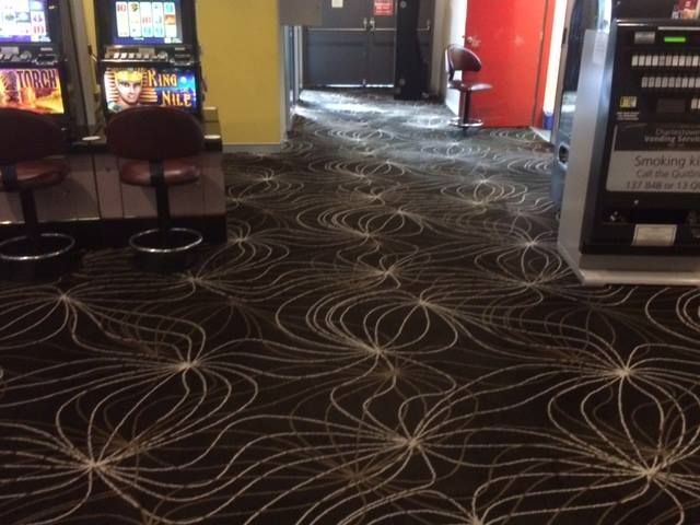 Indoor shot of a casino floor with black carpet featuring a swirling white line pattern, gaming machines, and stools.