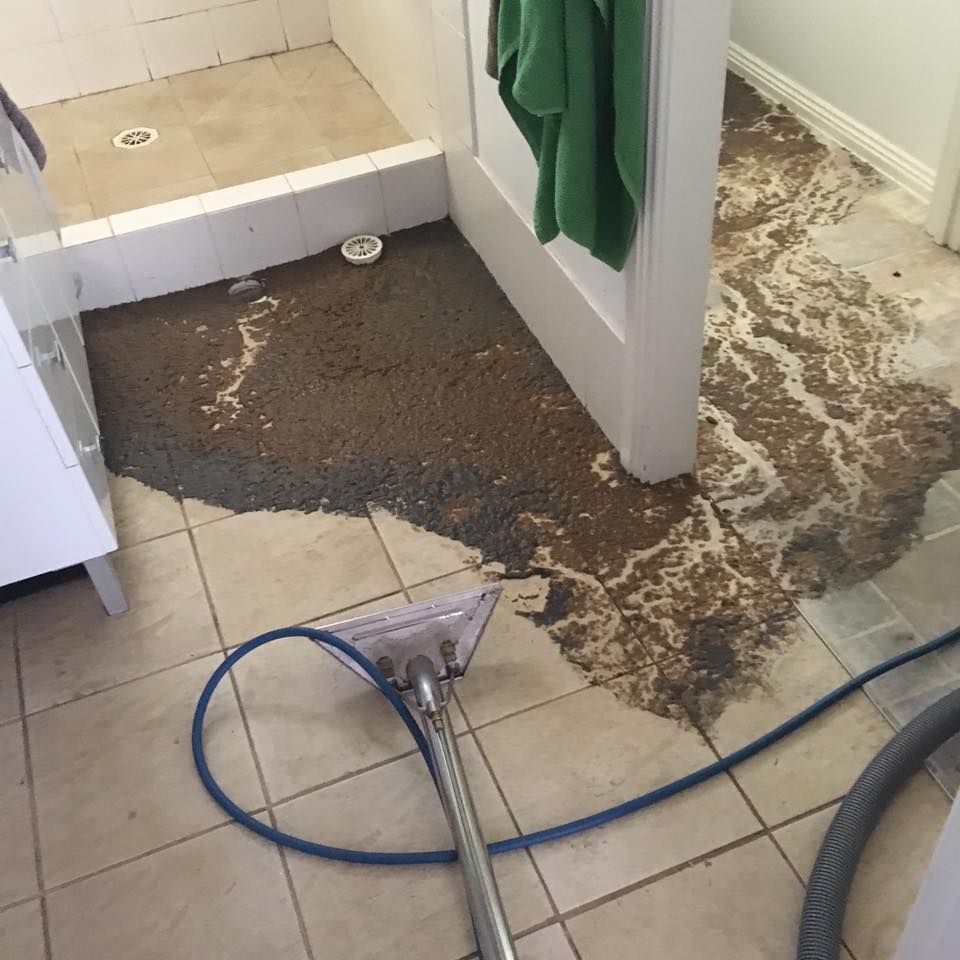 A carpet cleaning tool extracts dark, dirty water from tiled bathroom flooring near a shower and a doorway.