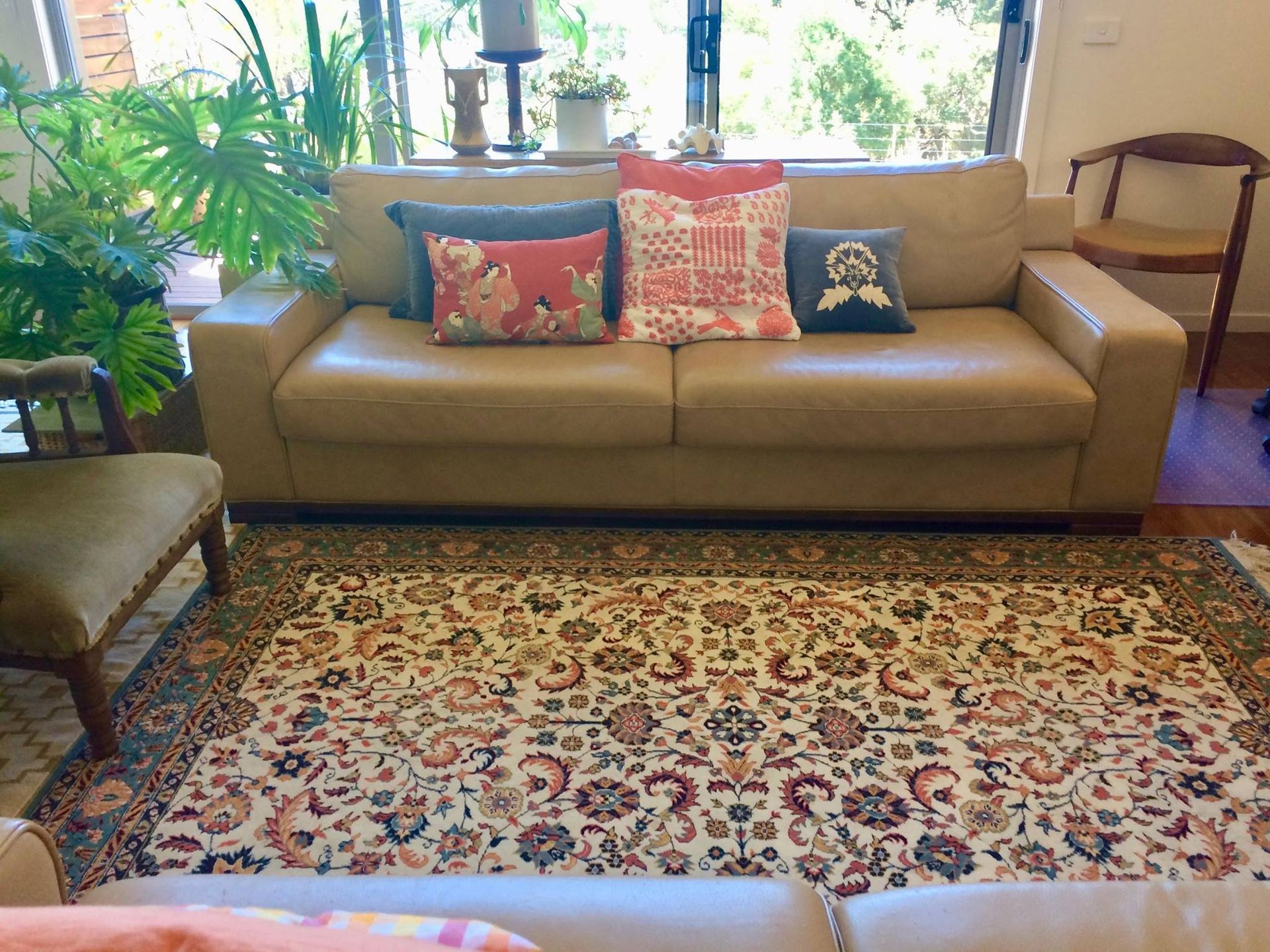 A beige couch with colorful throw pillows sits behind a patterned rug in a sunlit room with various plants and chairs.