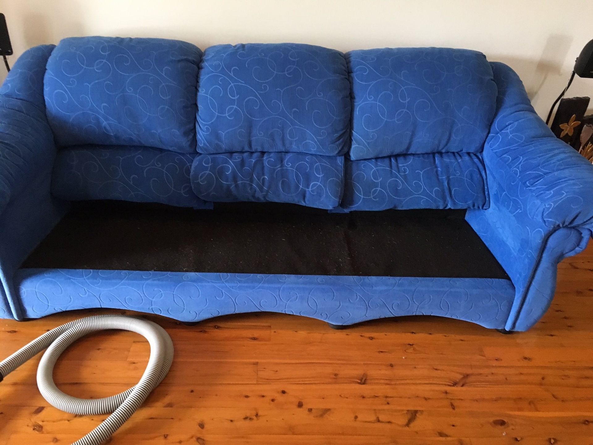 Blue three-cushion sofa with missing seat cushions, revealing a black base, sitting on a wooden floor.