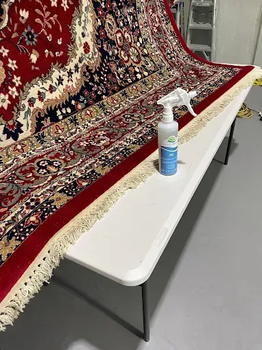A white spray bottle sits on a folding table next to a patterned red, cream, and black area rug.