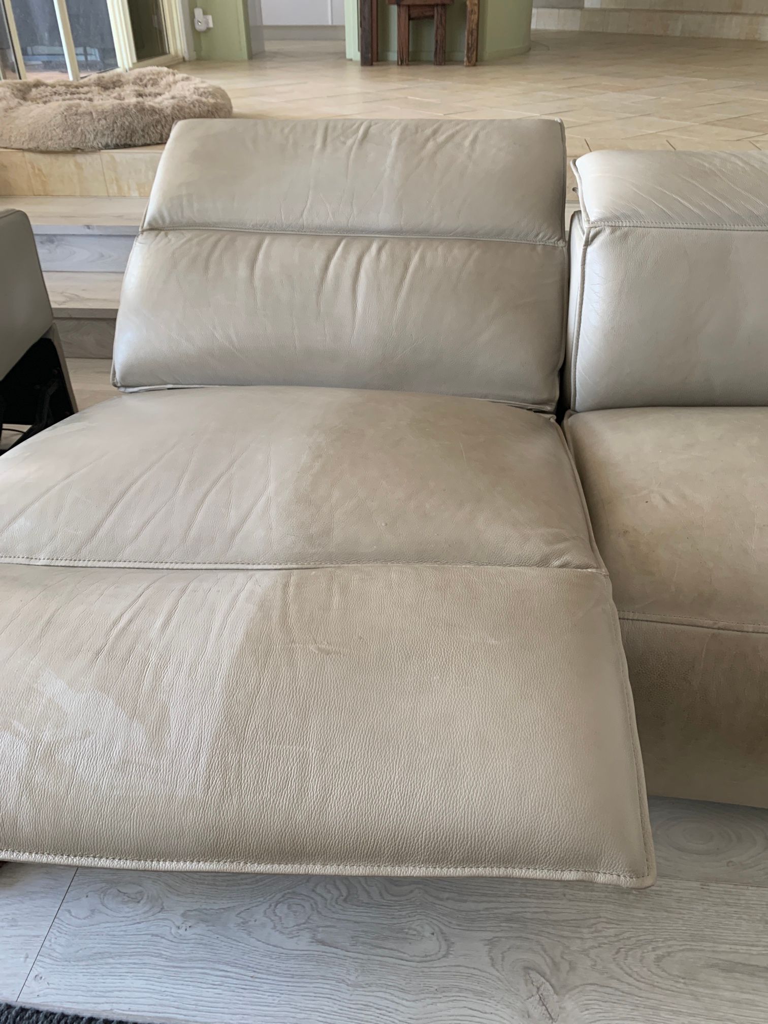 A beige leather recliner chair with a raised footrest, positioned on a light-colored tiled floor in a bright room.