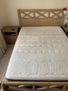 A light-wood bed frame with a crisscross headboard, fitted with a white tufted mattress, next to a matching nightstand.