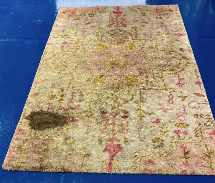 A rectangular area rug with a faded floral pattern in pink and gold, featuring a large dark soil stain on the bottom left.