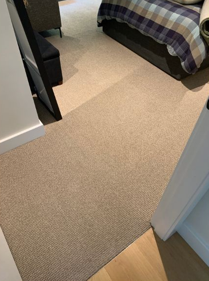 A view through a doorway into a bedroom with beige carpet, a bed with a plaid quilt, and a black storage bench.