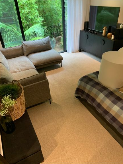 A living room with a beige sofa, a television on a black cabinet, and a large window looking out at green trees.