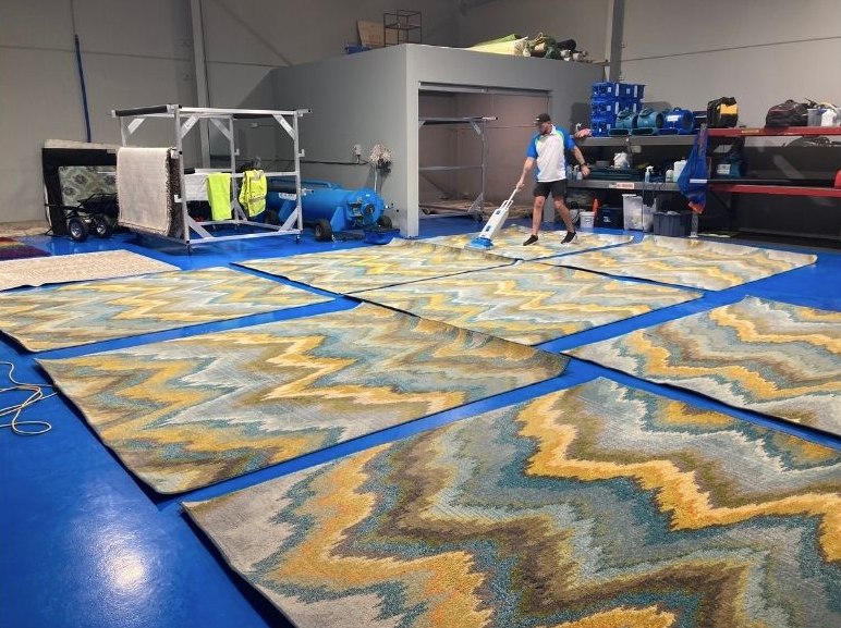 A person vacuums large area rugs with a blue, yellow, and white chevron pattern laid out on a blue warehouse floor.