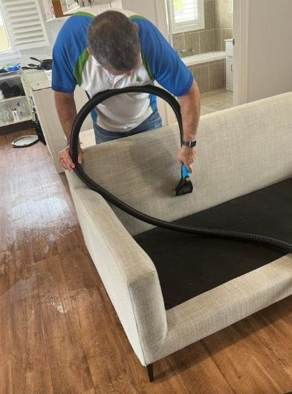 A person in a blue and white shirt deep-cleans a light-colored couch with a vacuum upholstery tool in a home setting.