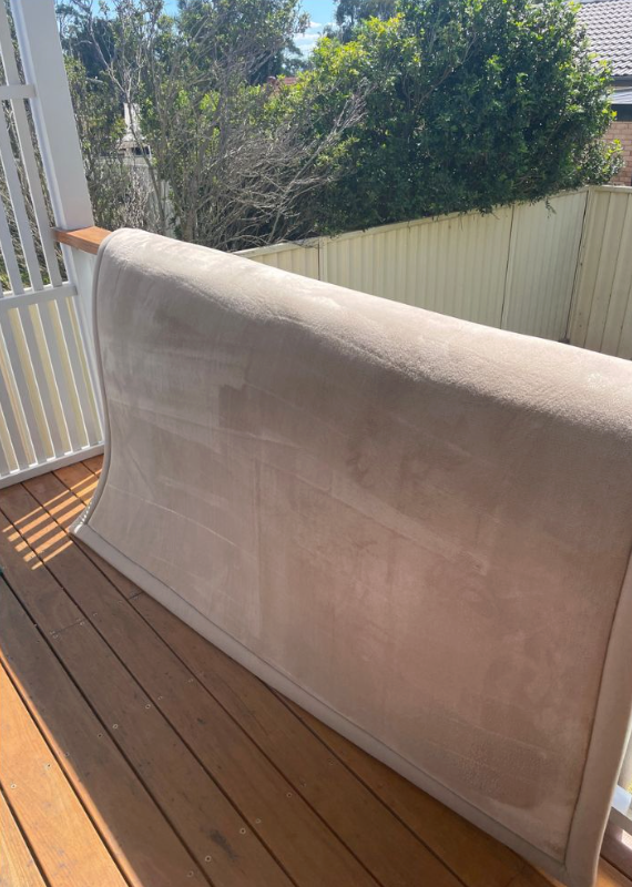 A large, beige, rectangular upholstered headboard leaning against a wooden deck railing outdoors.