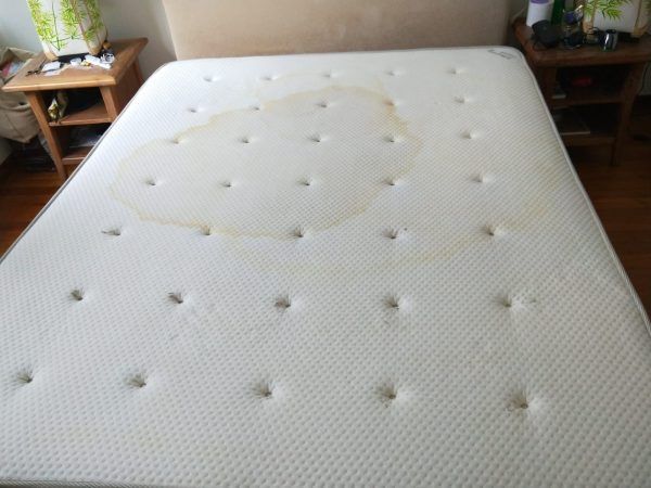 A mattress with a large, light brown organic stain on the surface, positioned between two bedside tables in a bedroom.