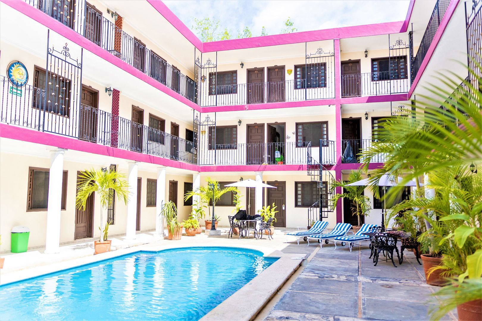 Patio del hotel con piscina, edificio rosa y blanco, palmeras, camastros y mesas.