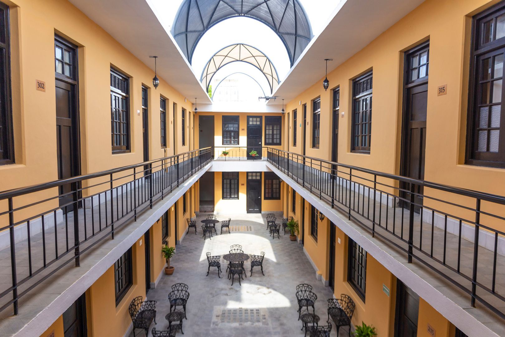 Patio interior con paredes amarillas, balcones, mesas y techo de cristal arqueado.
