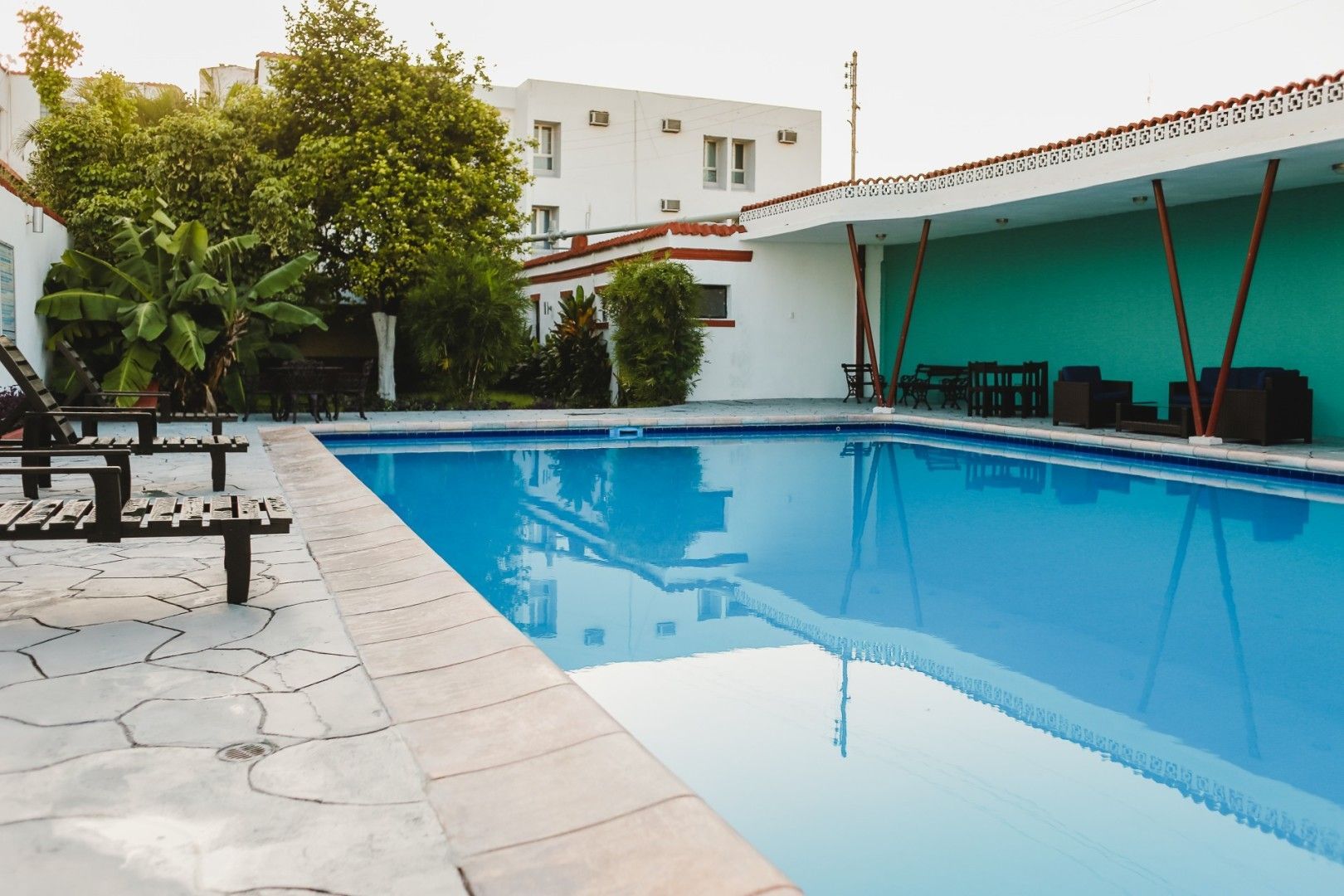 Piscina de agua azul brillante, rodeada de patio y edificio con árboles.
