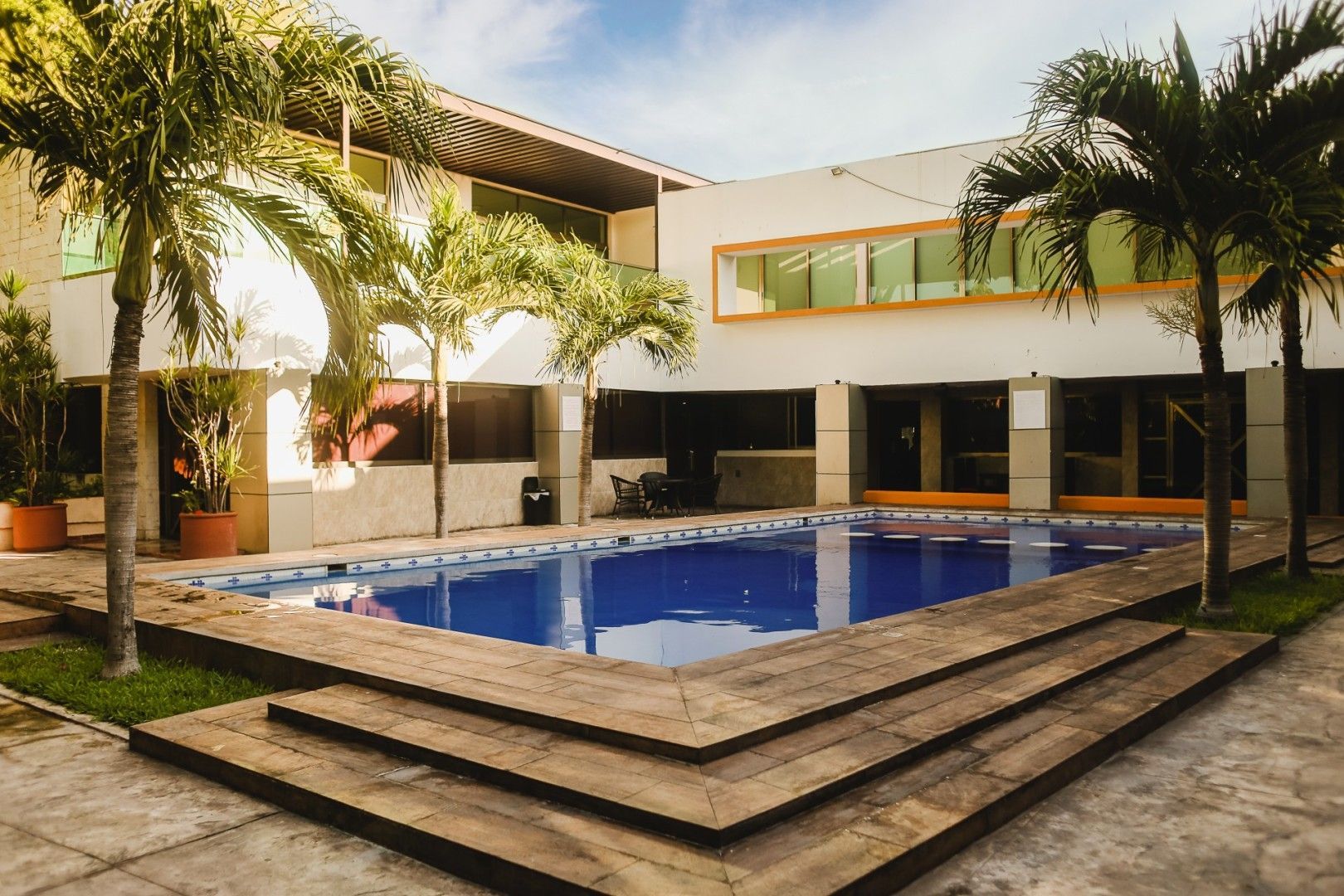 Piscina rodeada de un edificio con palmeras. Entorno soleado al aire libre.