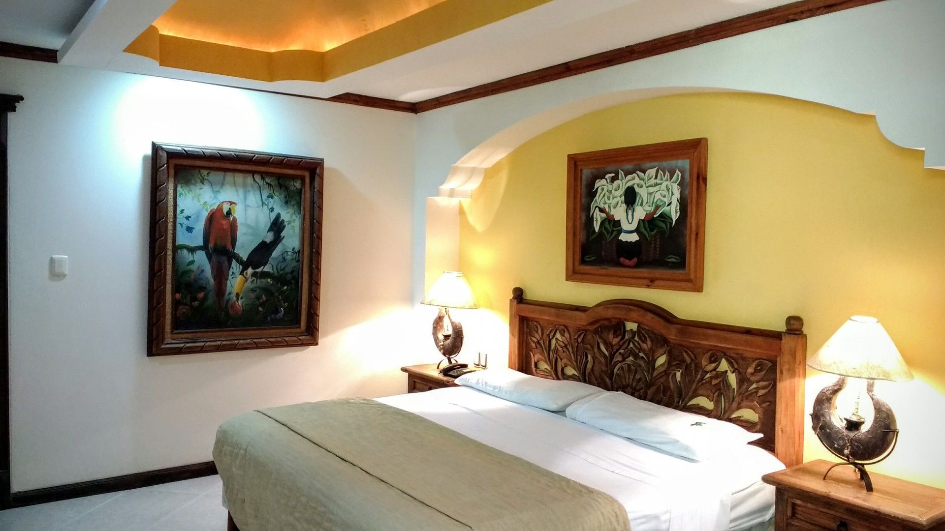 Dormitorio con cabecero de madera ornamentado, dos cuadros enmarcados y dos mesitas de noche con lámparas.