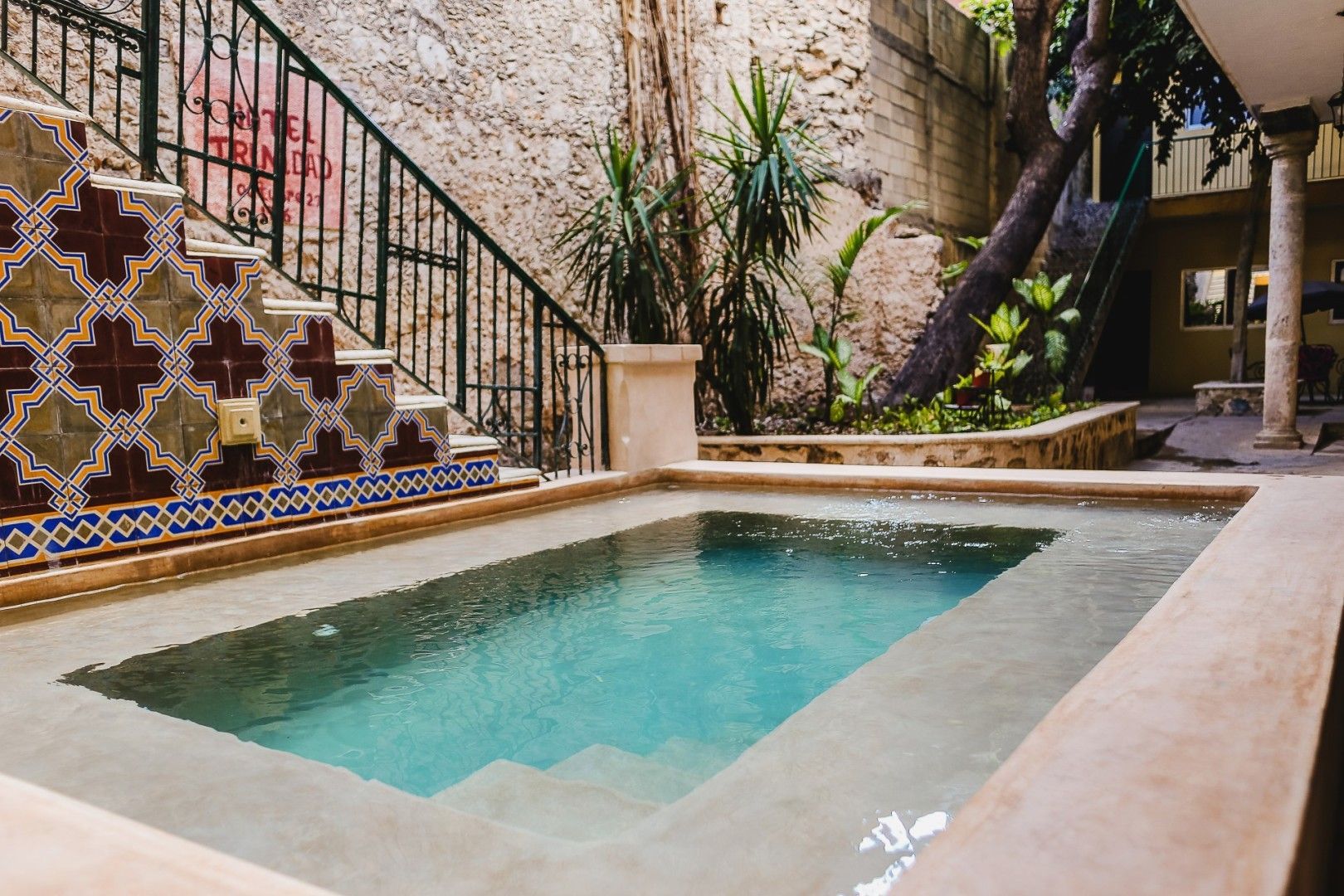 Pequeña piscina con agua turquesa, escalera de azulejos ornamentados, paredes de piedra y vegetación.