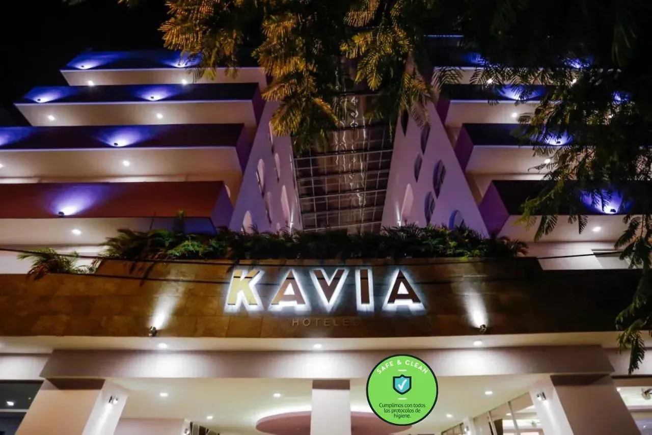 Entrada del hotel iluminada de noche. Letrero "KAVIA". Fachada morada y blanca.