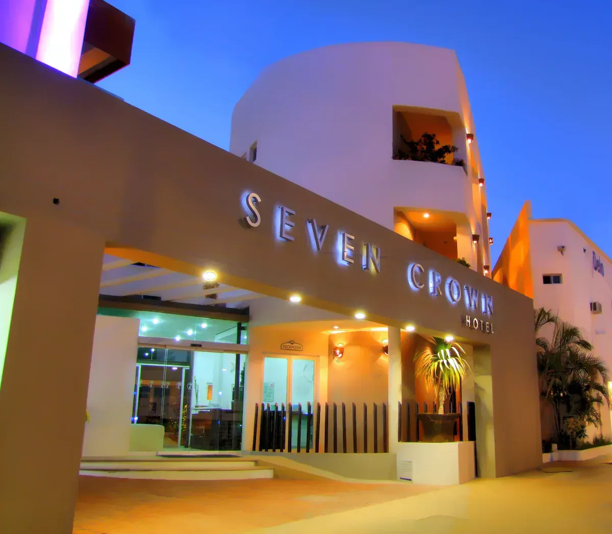 Entrada del Hotel Seven Crown al anochecer, cartel iluminado, edificio beige y blanco, palmera.