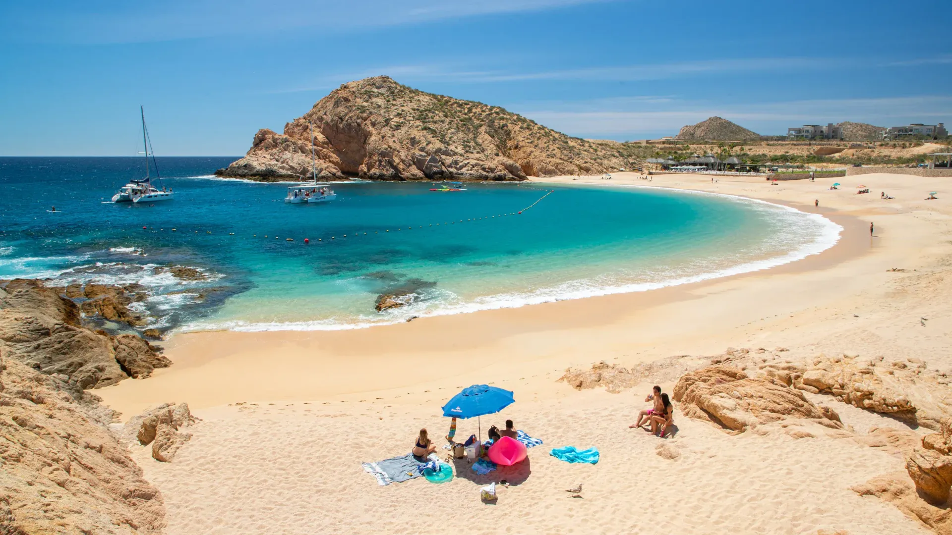 Playa de arena con agua turquesa, acantilados rocosos y algunas personas relajándose bajo una sombrilla azul.