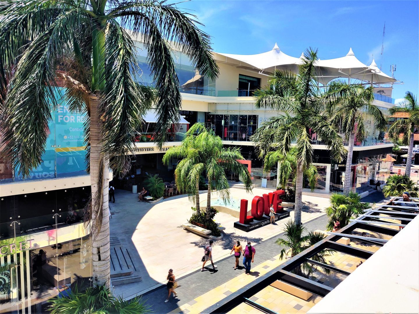 Un centro comercial soleado al aire libre con palmeras, una escultura roja brillante con la palabra "LOVE" y peatones.