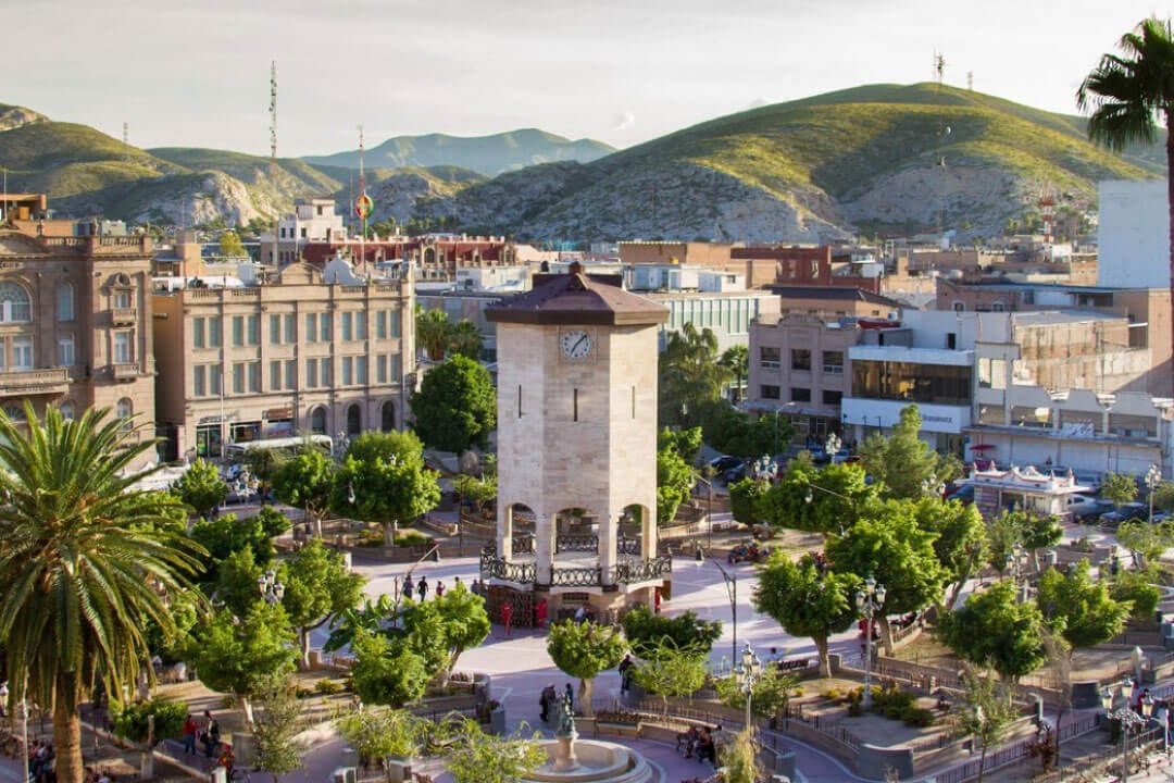 Plaza de la ciudad con torre del reloj, árboles, edificios y colinas al fondo.