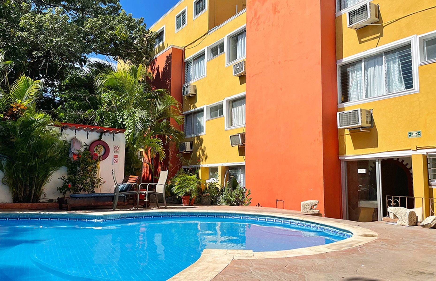 Piscina frente a un hotel de colores amarillo y naranja. Palmeras y vegetación.