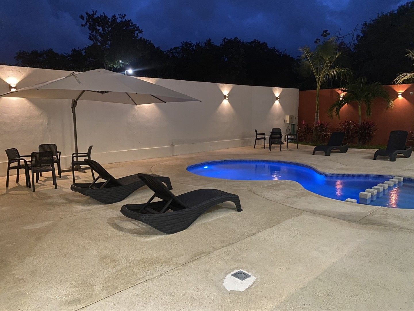 Área de piscina por la noche con tumbonas, sombrillas, mesas e iluminación ambiental contra una pared blanca.