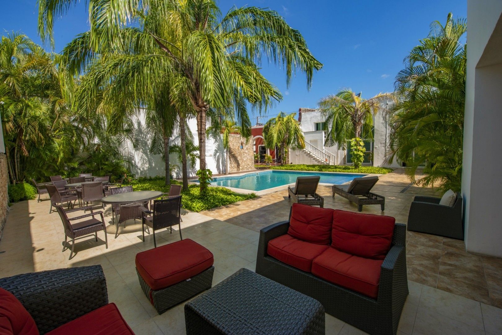 Patio exterior con piscina. Sofás rojos, sillas, mesas y palmeras. Cielo azul y soleado.