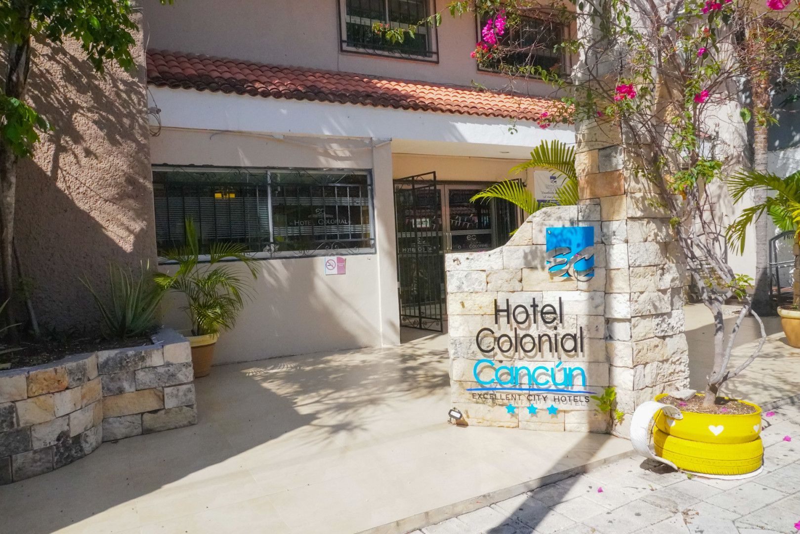 Entrada del Hotel Colonial Cancún. Edificio blanco con letrero, puerta, detalles de piedra y concreto, y plantas.