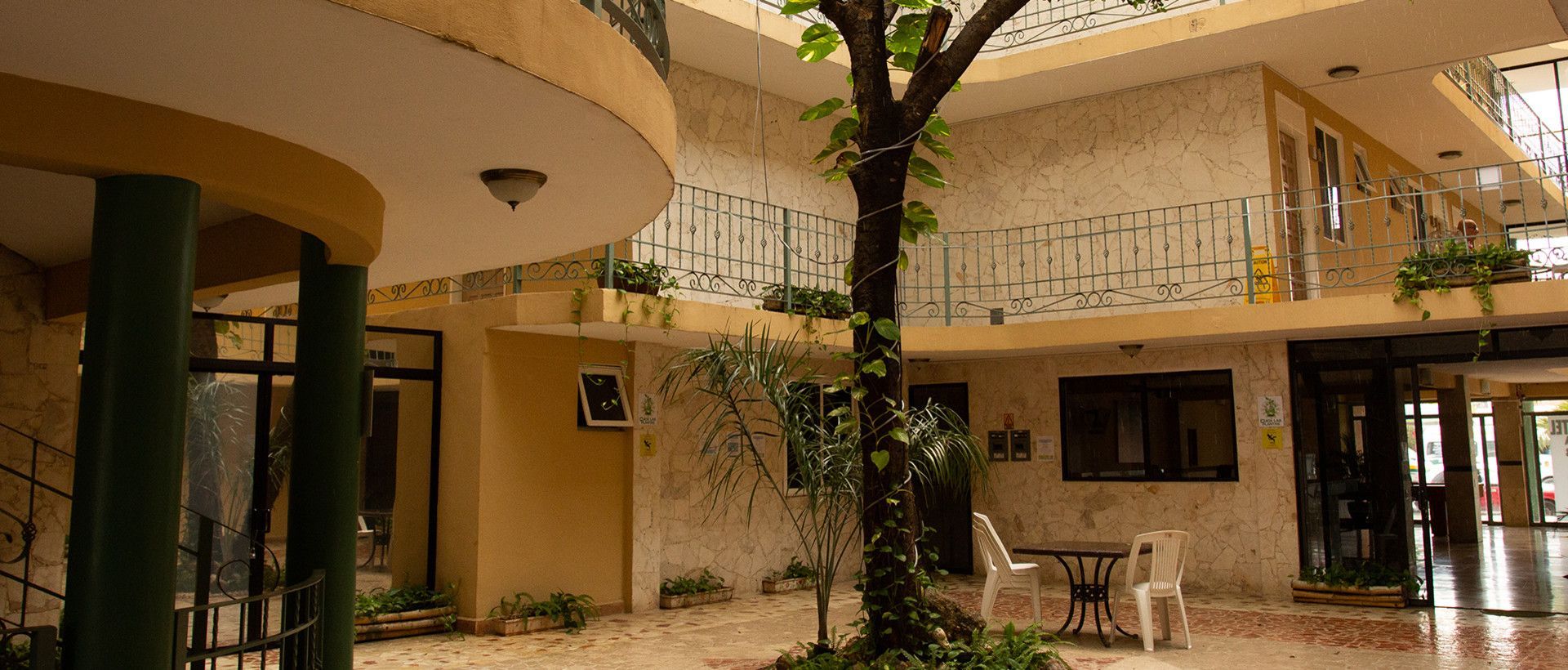 Patio con edificio de paredes amarillas, árbol, plantas pequeñas y mesa y sillas blancas.