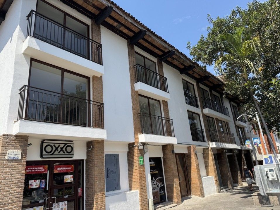 Edificio con tienda OXXO, balcones y árboles. Exterior blanco y ladrillo. Día soleado.