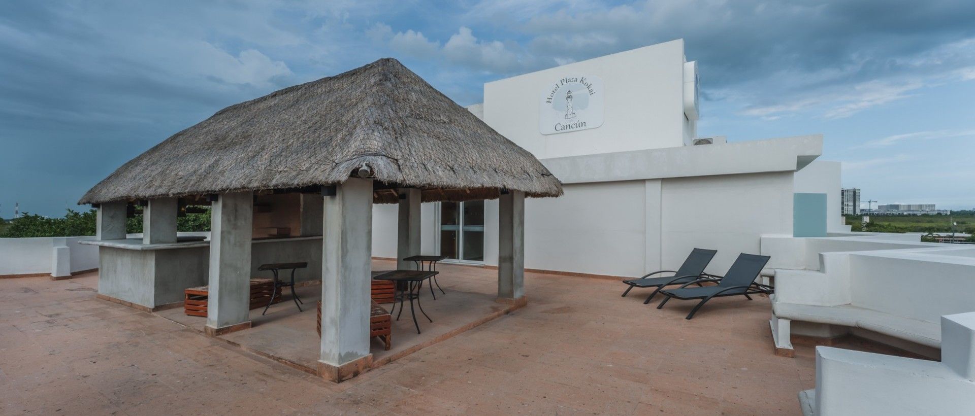Azotea con un refugio con techo de paja, sillas y un edificio blanco bajo un cielo nublado.
