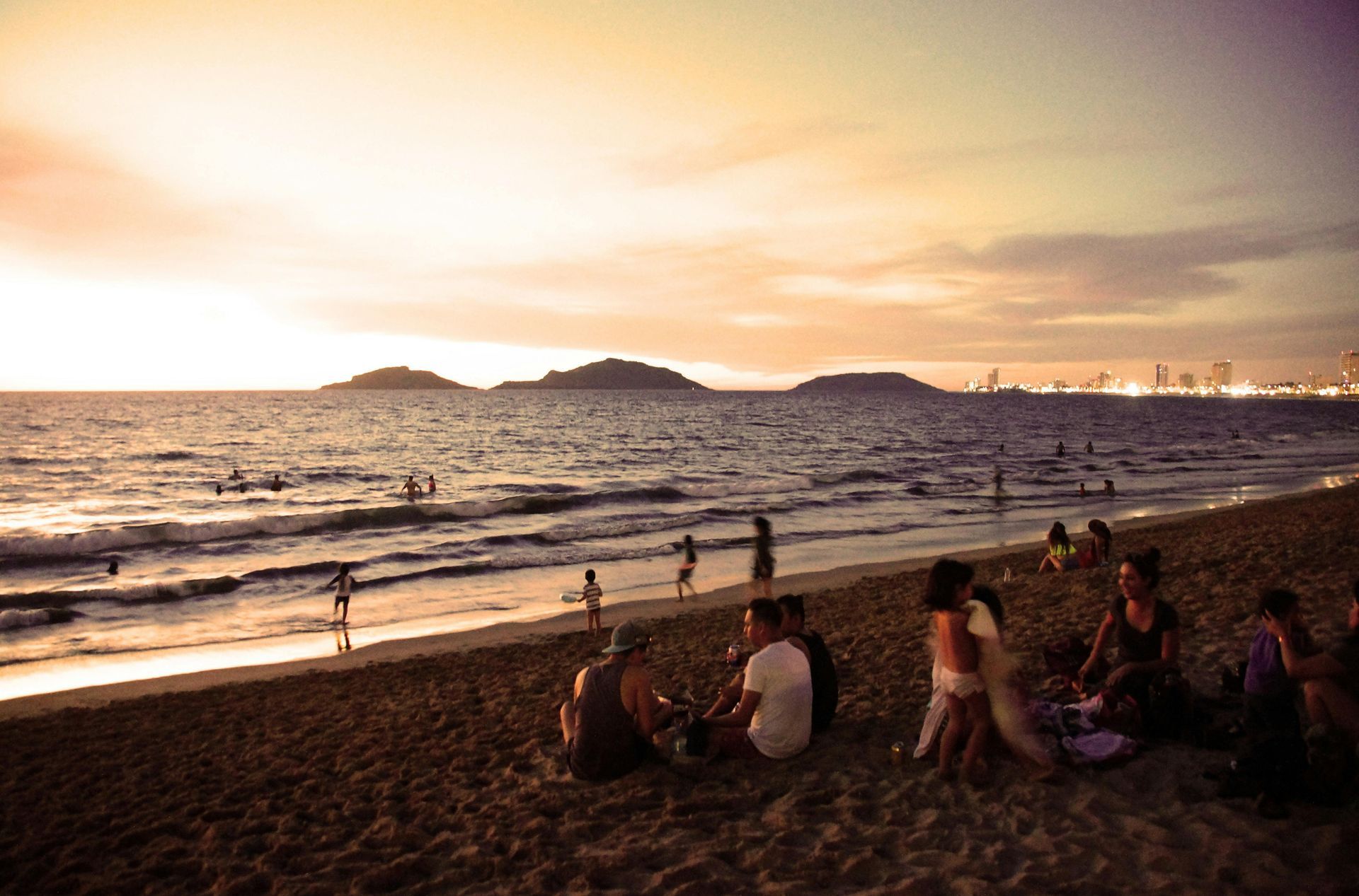 Escena de playa al atardecer, gente relajándose en la arena, olas, islas en el horizonte, luz dorada.