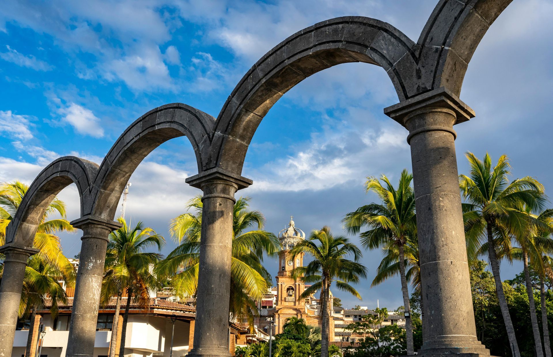 Arcos de piedra enmarcan una vista de una iglesia y palmeras contra un cielo azul nublado.