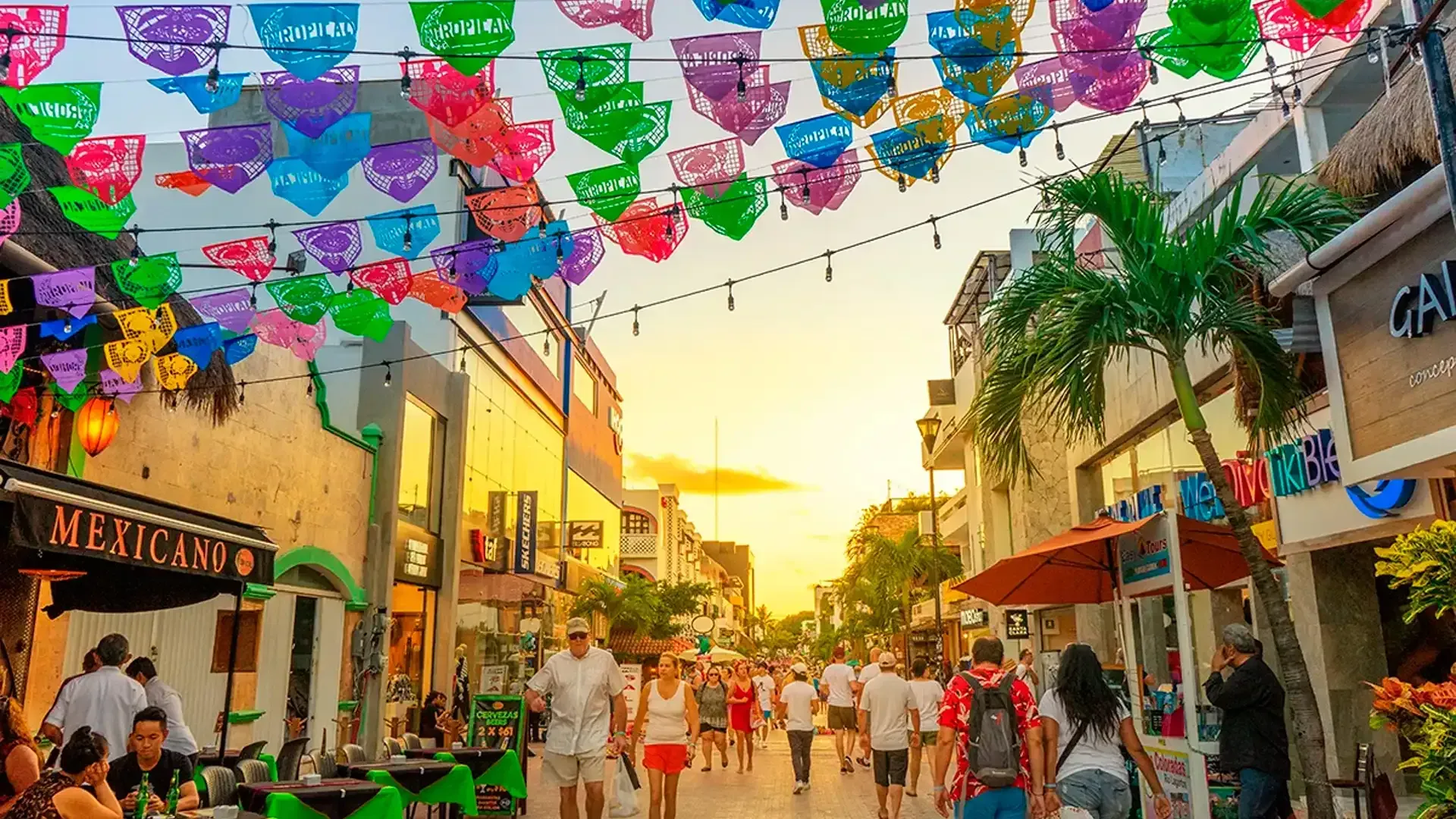 Gente caminando por una calle llena de tiendas, bajo coloridas pancartas de papel y luces de cadena, al atardecer.