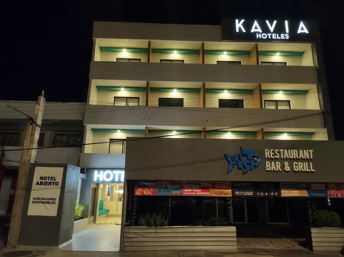 Edificio de Kavia Hoteles de noche con habitaciones iluminadas, restaurante adyacente y cartel de "Hotel Abierto".