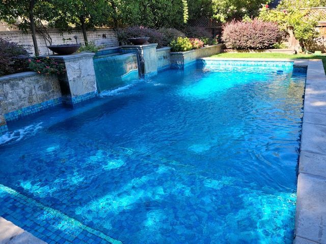 Blue tiled swimming pool with waterfall features, set in a sunny backyard with greenery.