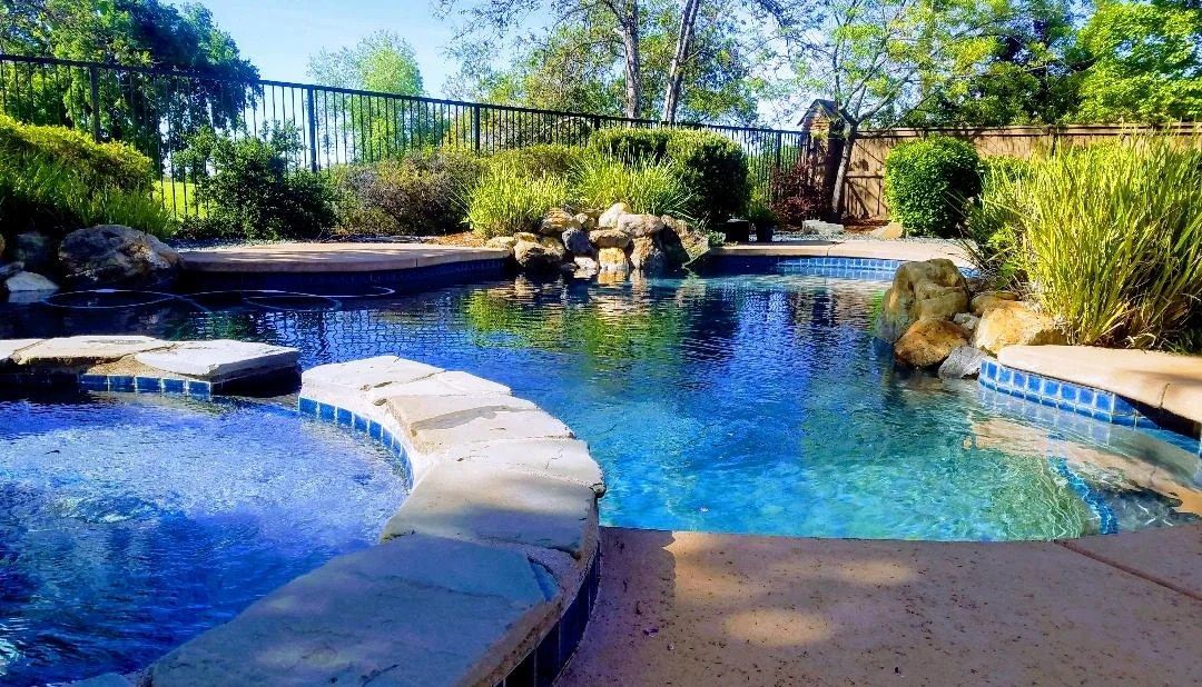 Swimming pool with blue water and stone surround, surrounded by lush greenery and trees.