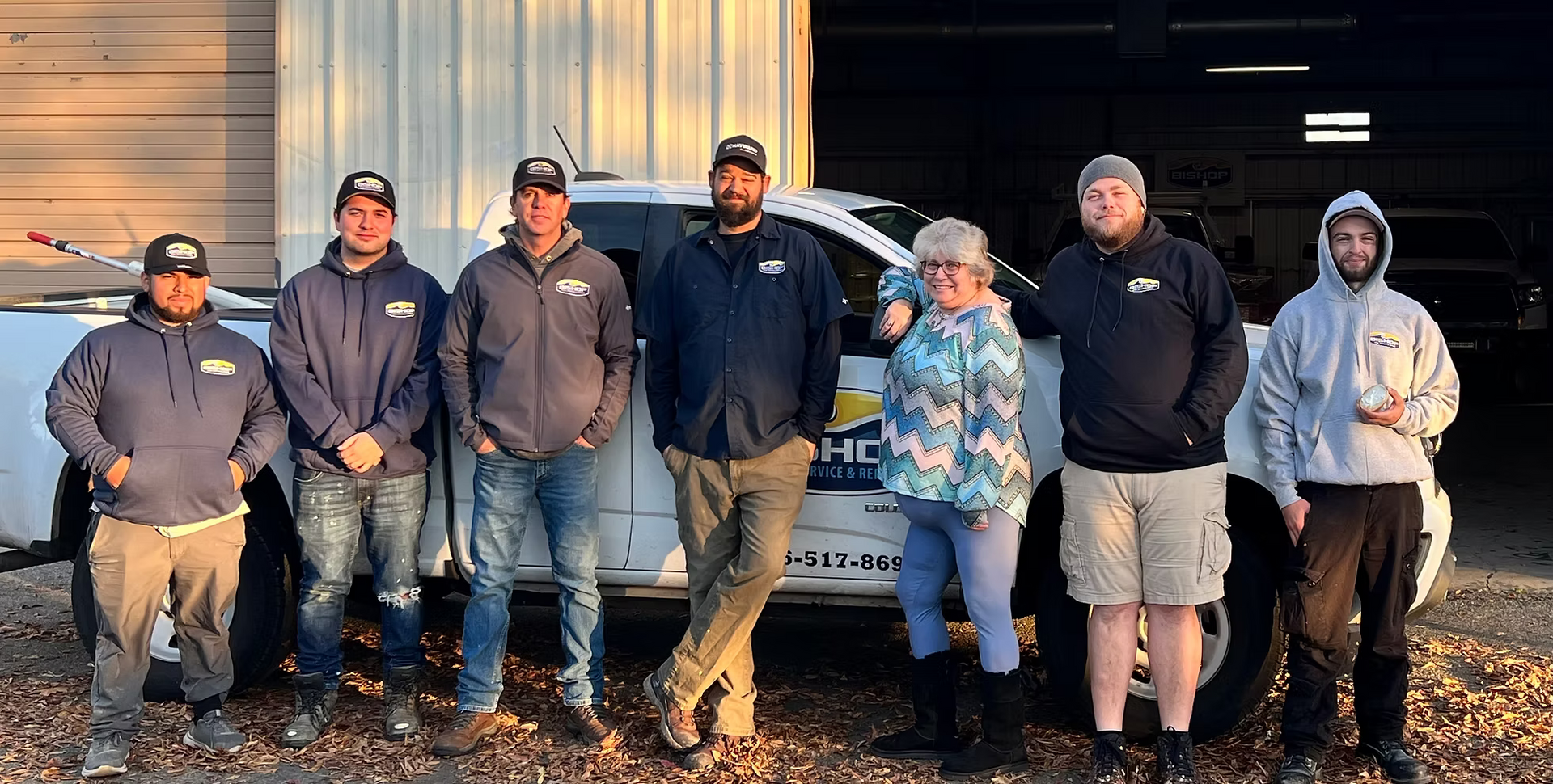 Group of people standing in front of a white truck.  They are smiling. Outdoors, sunny.