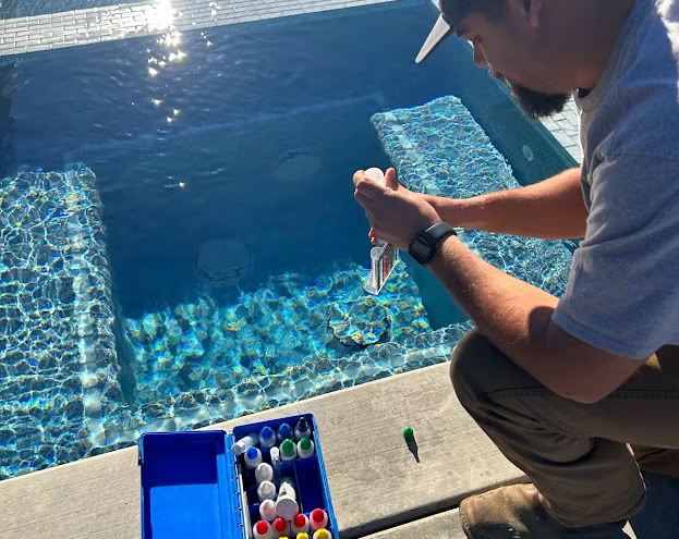 Man testing pool water with a kit near a blue-tiled pool, under bright sunlight.