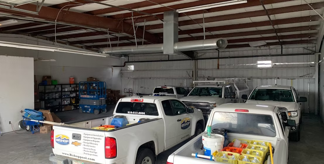 White pickup trucks parked inside a garage. Shelves, tools, and ductwork are visible.