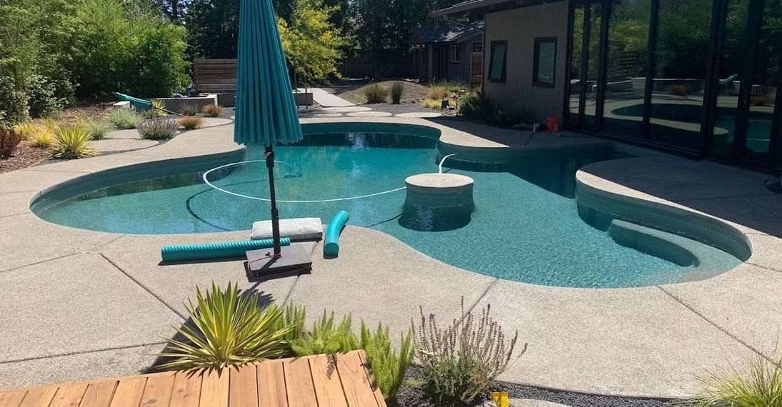 Pool with turquoise water surrounded by concrete patio and landscaping, patio umbrella open.