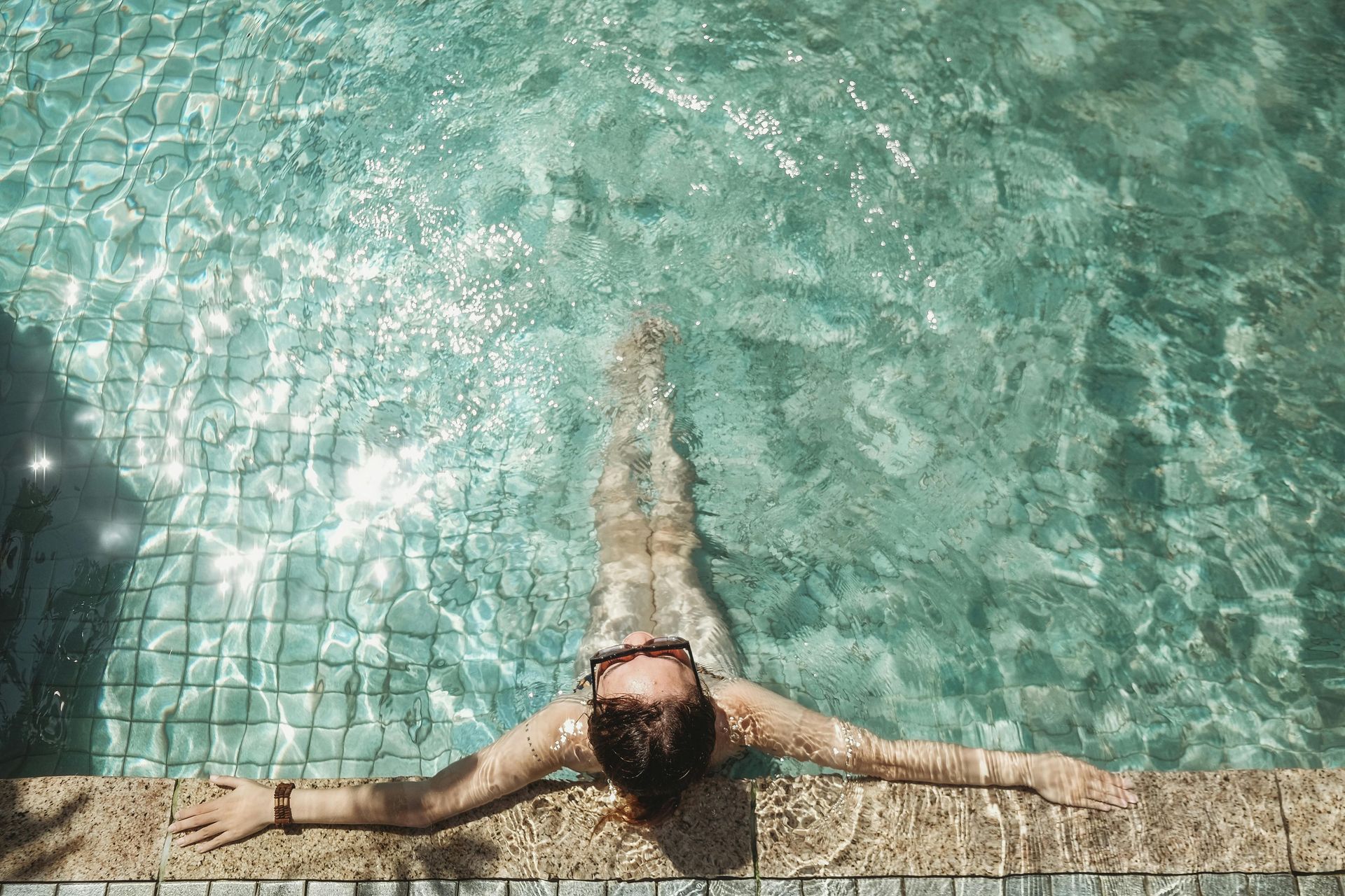 Person relaxing in a pool, arms outstretched. Sunlight reflects on the clear water.