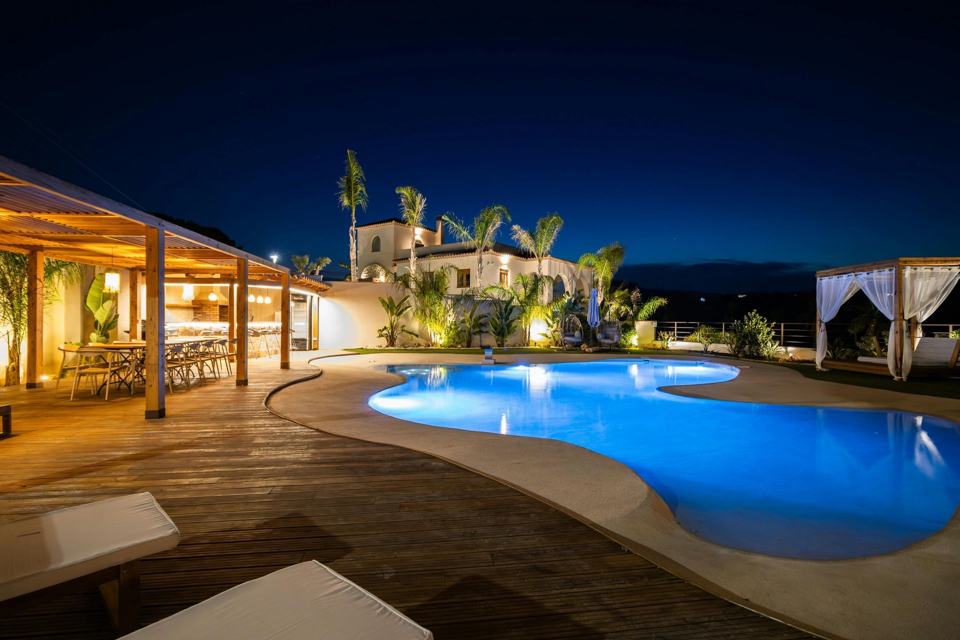 Nighttime view of a pool with glowing blue water, outdoor dining, and a white villa in the background.