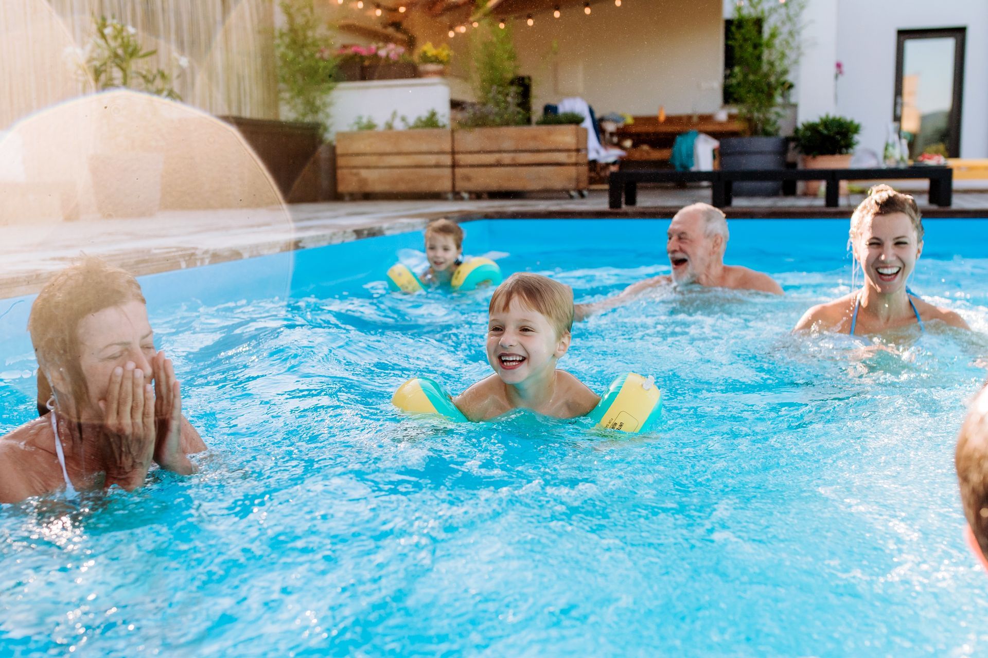 Family in a pool, smiling and laughing. Two children with arm floaties. Sunlit outdoor setting.