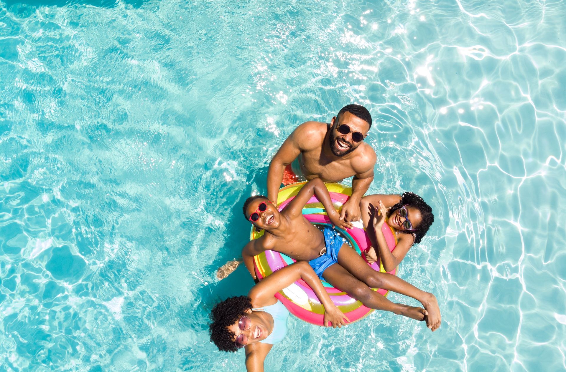Family in a pool on inflatable rings, smiling. Clear blue water, sunny day.